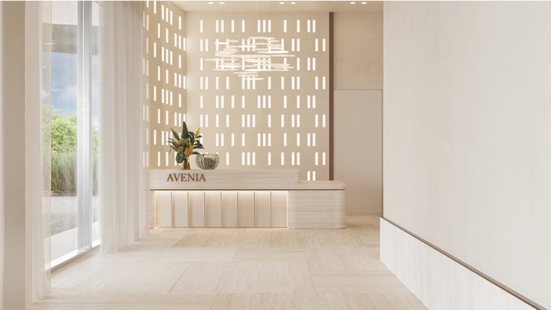 Avenia Aventura. A minimal, modern lobby with neutral tones, a plant on the reception desk labeled AVENIA, and patterned light filtering in.
