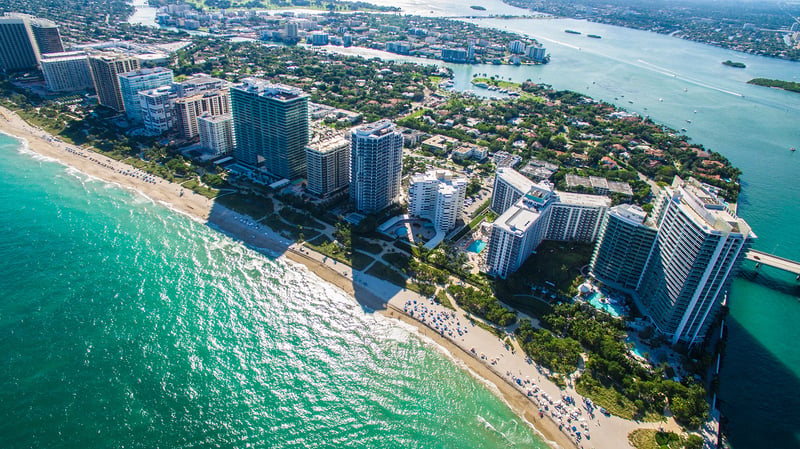 Aerial view of tall beachfront buildings along a sandy coastline with turquoise water and a city in the background. Featuring Bal Harbour, coastal, and skyline.