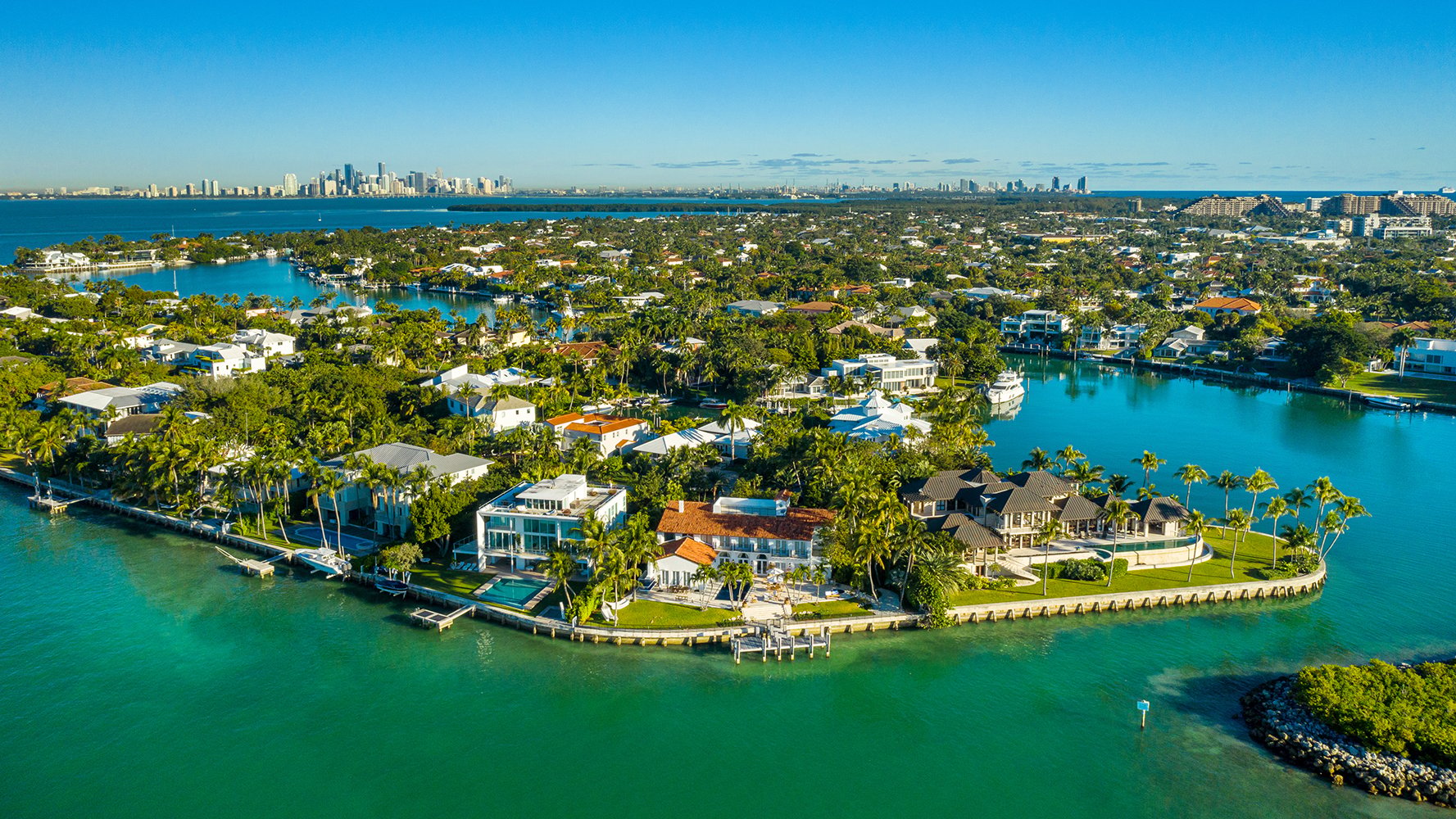 Aerial view of a waterfront neighborhood with luxury homes, boats, palm trees, and turquoise water, with a city skyline in the background. Featuring Miami.