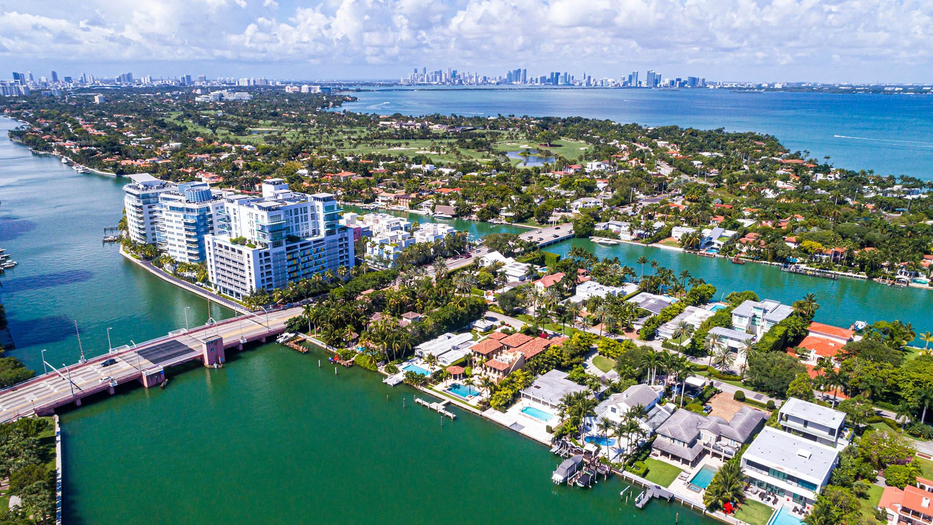 Aerial waterfront view of Allison Island in Miami Beach showing luxury and ultra luxury condos, waterfront homes, canals, a bridge, lush island streets, Biscayne Bay, and the distant downtown Miami skyline.