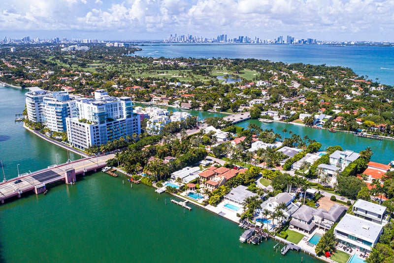 Aerial waterfront view of Allison Island in Miami Beach showing luxury and ultra luxury condos, waterfront homes, canals, a bridge, lush island streets, Biscayne Bay, and the distant downtown Miami skyline.