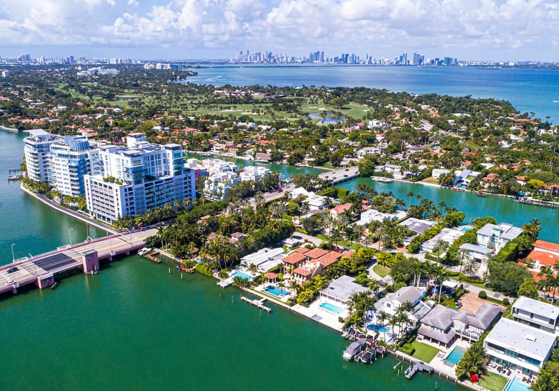 Aerial waterfront view of Allison Island in Miami Beach showing luxury and ultra luxury condos, waterfront homes, canals, a bridge, lush island streets, Biscayne Bay, and the distant downtown Miami skyline.
