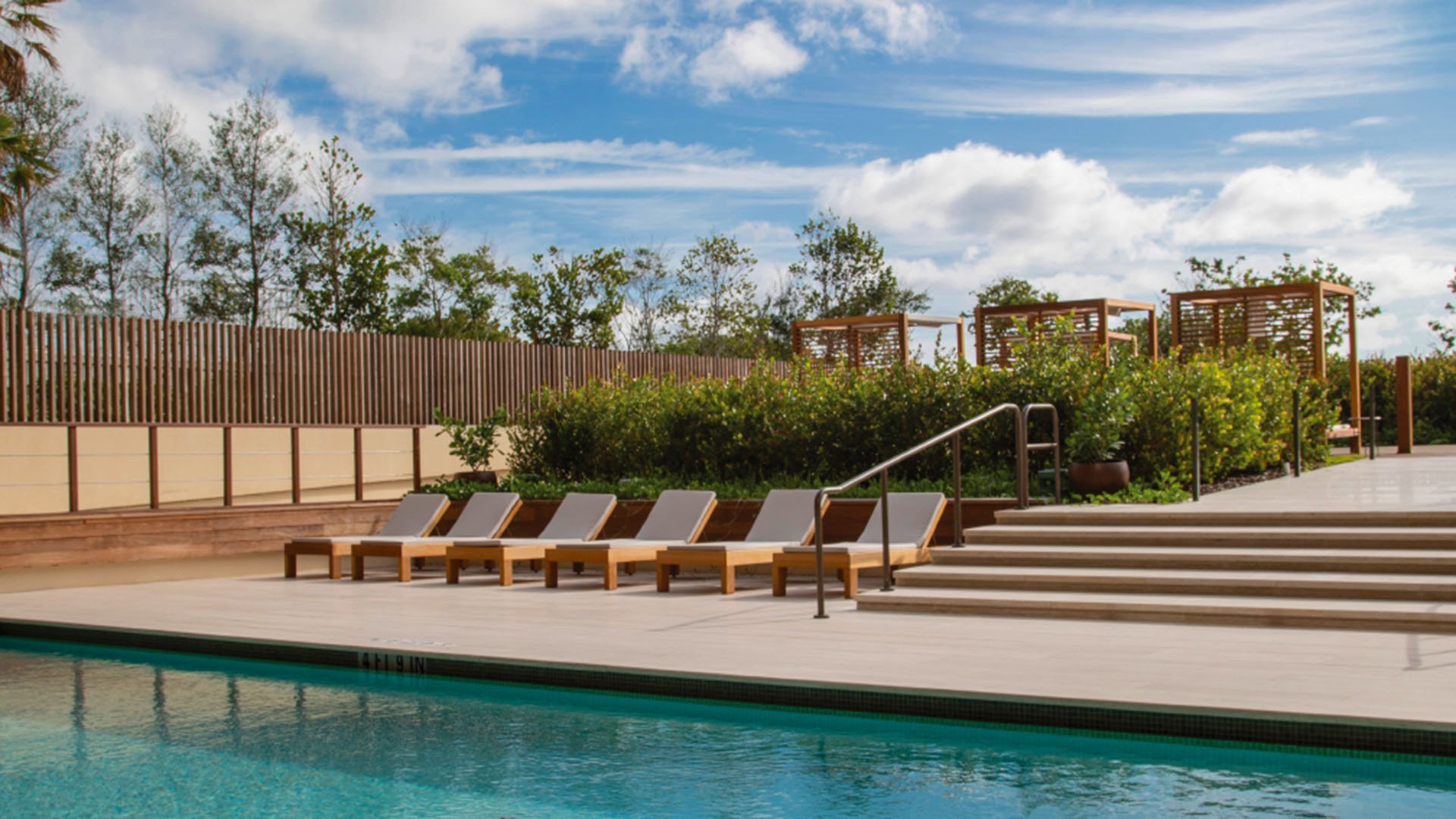 Outdoor pool deck at Arte Surfside, Surfside, Florida, showing luxury and ultra luxury condos with sun loungers, manicured hedges, broad steps, and pergola lounge areas beside the water.