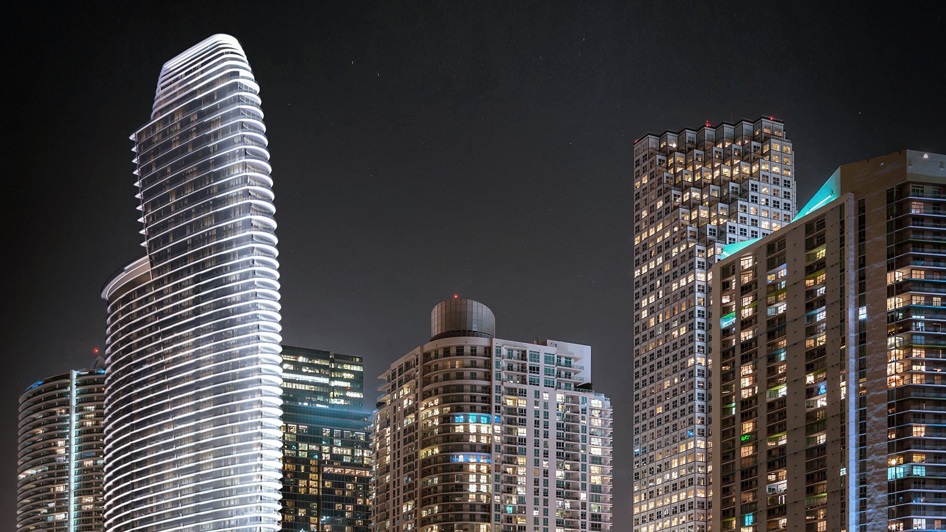 Aston Martin Residences in Downtown Miami luxury and ultra luxury condos nighttime skyline closeup with the illuminated curved tower rising among neighboring high-rises.