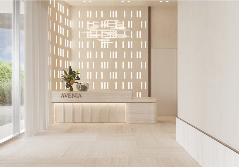 Avenia Aventura. A minimal, modern lobby with neutral tones, a plant on the reception desk labeled AVENIA, and patterned light filtering in.