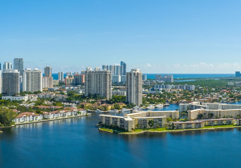 Aventura luxury and ultra luxury condos in an aerial waterfront view with high-rise residential towers, calm intracoastal canals, low-rise buildings, boats, and the ocean horizon under a clear blue sky.