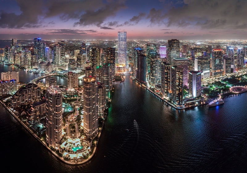 Baccarat Residences in Brickell, Miami, luxury and ultra luxury condos featuring a twilight aerial of the riverfront skyline, illuminated towers, waterfront streets, and glowing city lights.