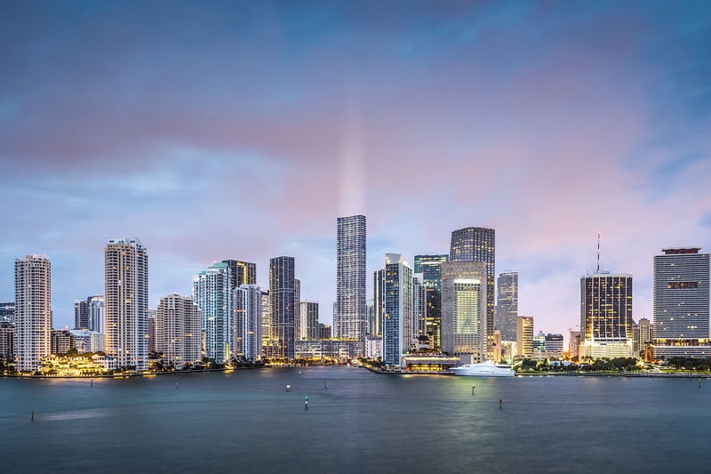 Baccarat Residences in Brickell, Miami, luxury and ultra luxury condos featuring an evening panorama of the waterfront skyline, illuminated towers, soft clouds, and calm bay water in the foreground.