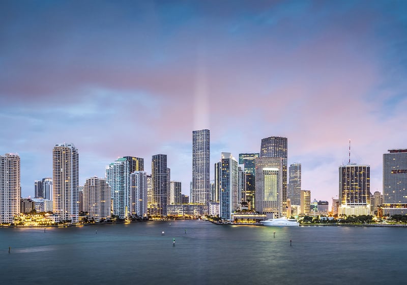 Baccarat Residences in Brickell, Miami, luxury and ultra luxury condos featuring an evening panorama of the waterfront skyline, illuminated towers, soft clouds, and calm bay water in the foreground.