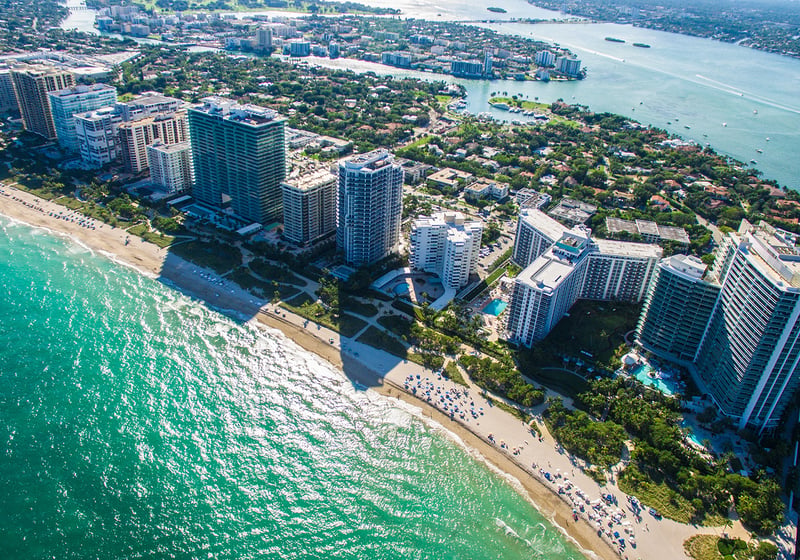 Aerial view of tall beachfront buildings along a sandy coastline with turquoise water and a city in the background. Featuring Bal Harbour, coastal, and skyline.