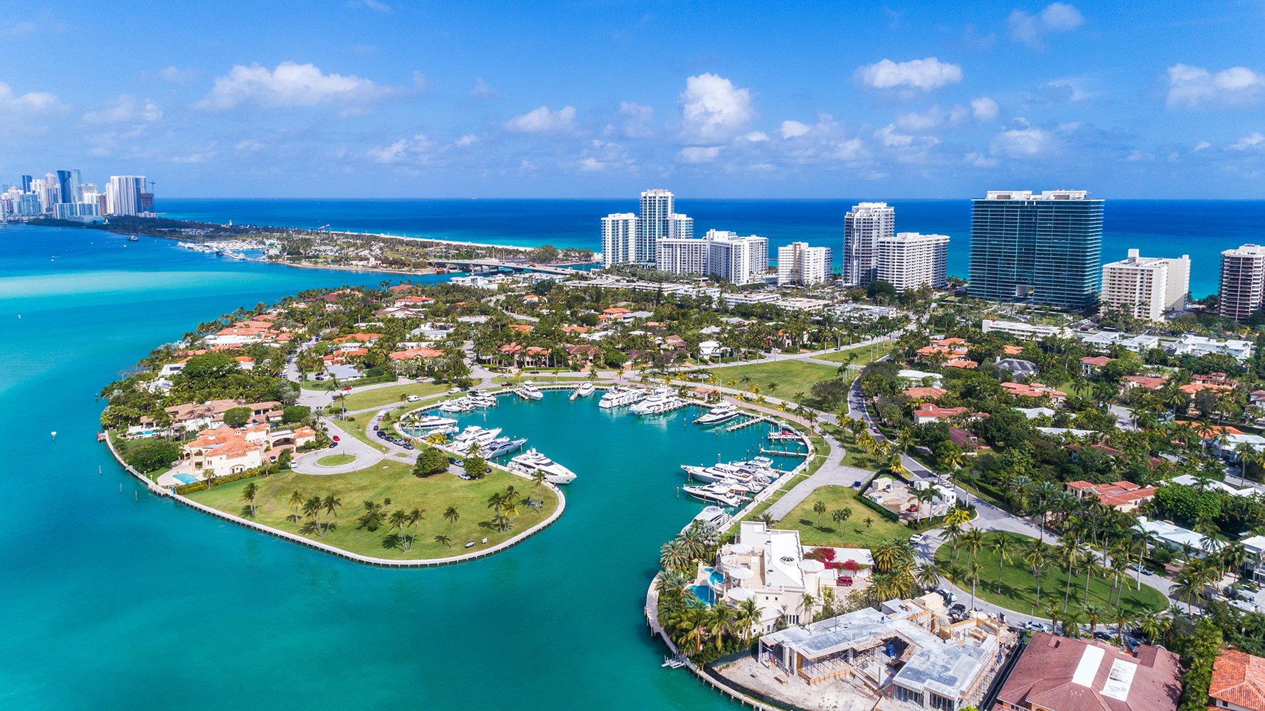 Aerial view of a coastal city with turquoise water, boats in a marina, palm trees, and high-rise buildings in the background. Featuring Bal Harbour and Miami skyline.