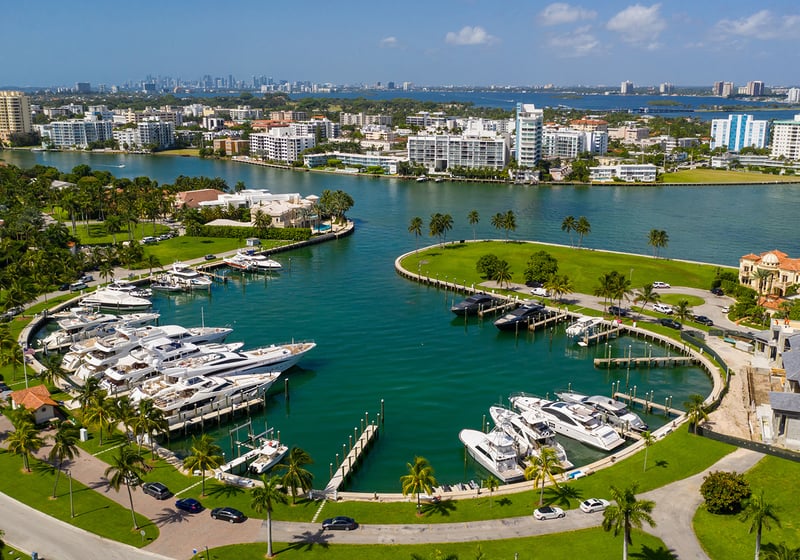 Aerial view of a marina with yachts, surrounded by water, palm trees, and modern buildings under a blue sky. Featuring Bal Harbour, waterfront, and Miami skyline.