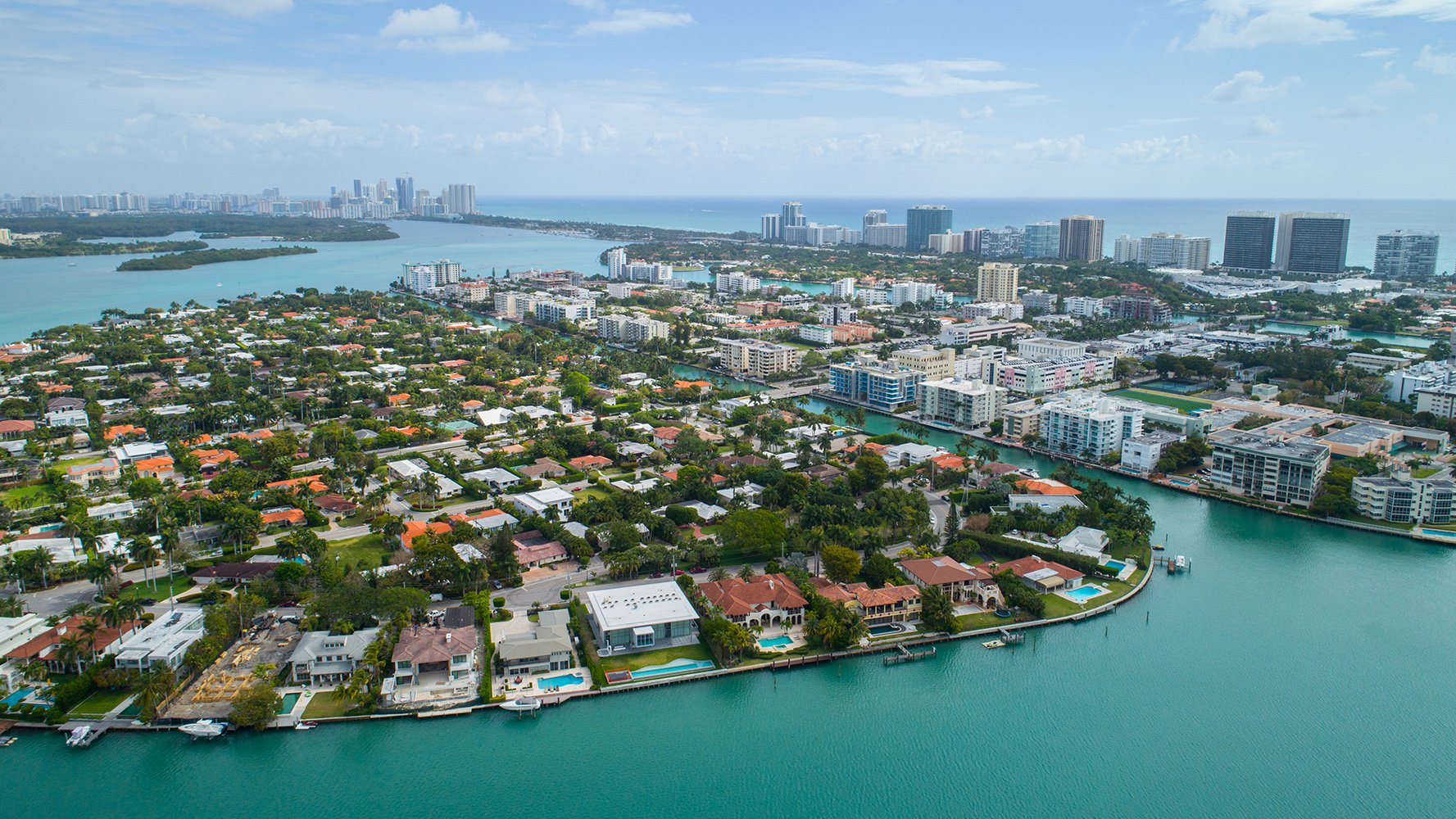 Aerial view of a coastal city with residential homes, waterways, and high-rise buildings under a partly cloudy sky. Featuring Bay, Harbor, and Miami.