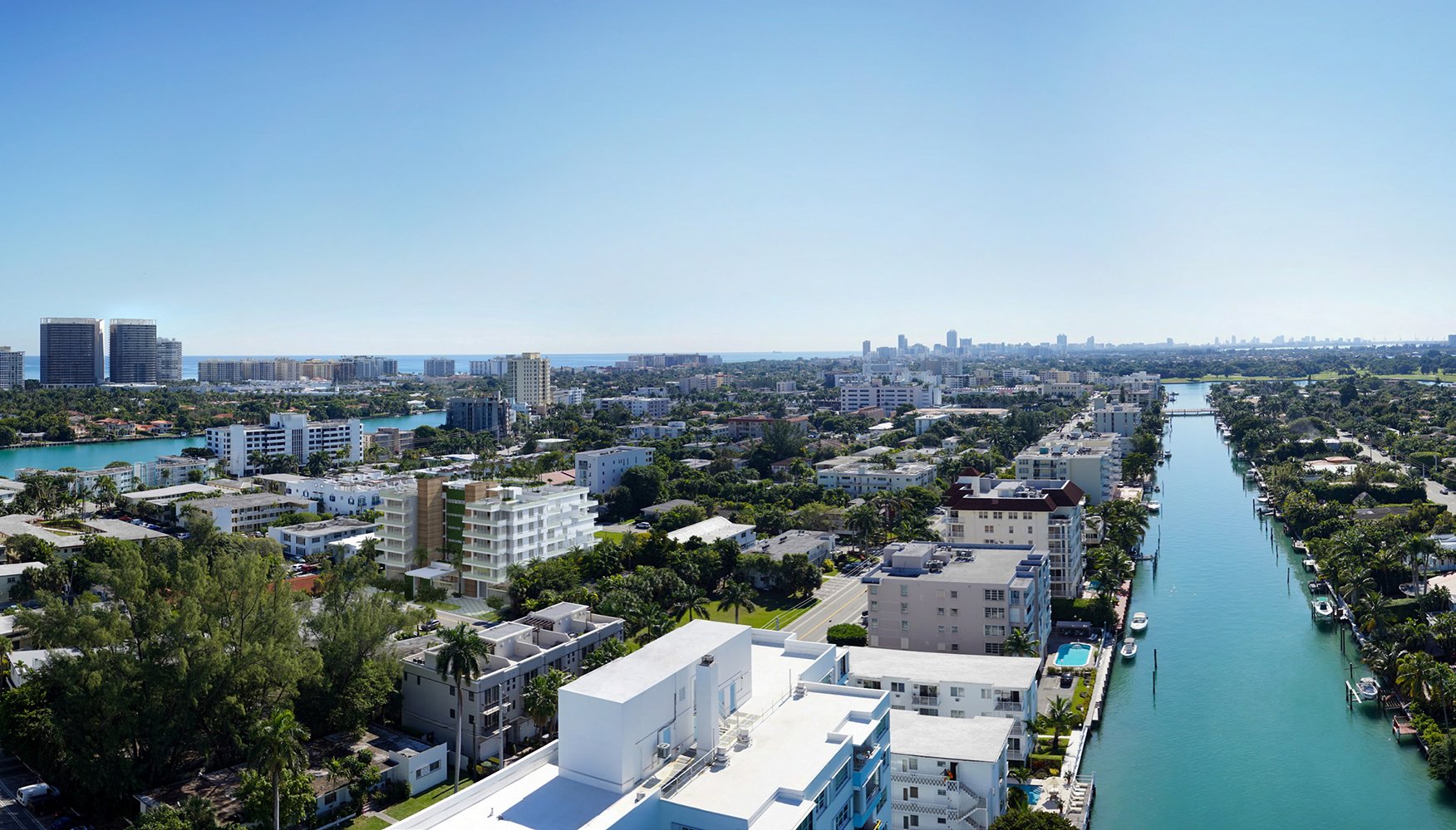 Aerial view of Miami with buildings, a canal lined with boats, and the ocean on the horizon under a clear blue sky. Featuring Bay, Harbor, cityscape, and waterfront.