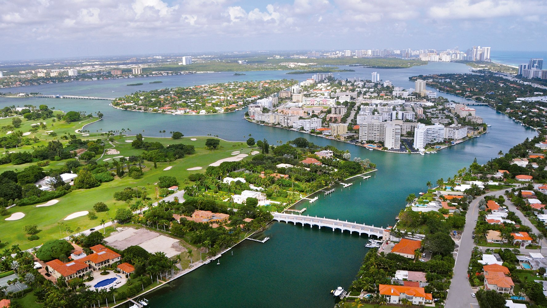 Aerial view of a coastal city with green golf courses, islands, bridges, and high-rise buildings surrounding blue waterways. Featuring Bay, Harbor, Miami, and homes.