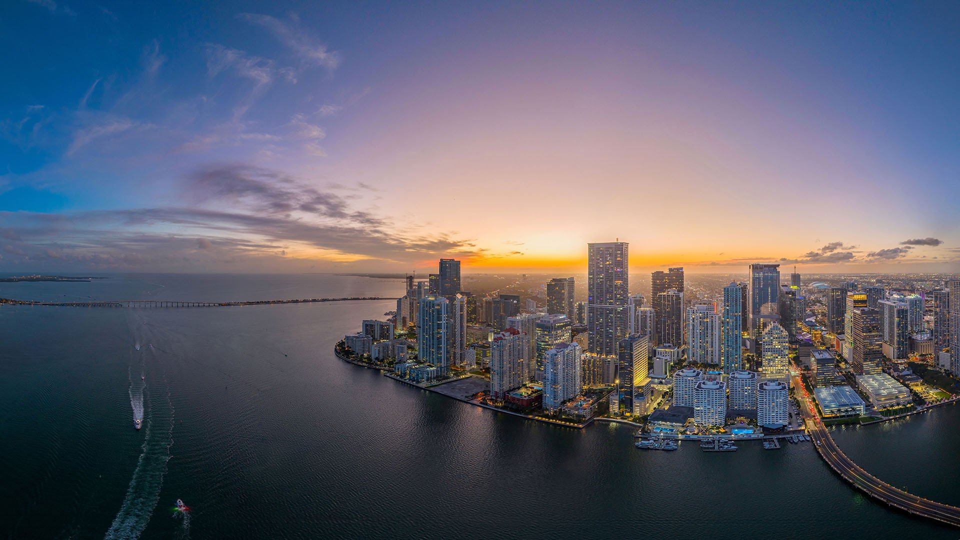 Wide sunset aerial of Downtown Miami along Biscayne Bay with boat wakes and horizon glow, showcasing luxury and ultra luxury condos with preconstruction and resale options near Brickell Key, Miami, Florida.