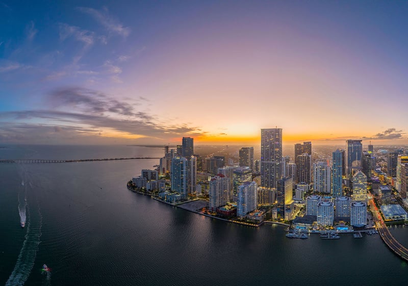 Wide sunset aerial of Downtown Miami along Biscayne Bay with boat wakes and horizon glow, showcasing luxury and ultra luxury condos with preconstruction and resale options near Brickell Key, Miami, Florida.
