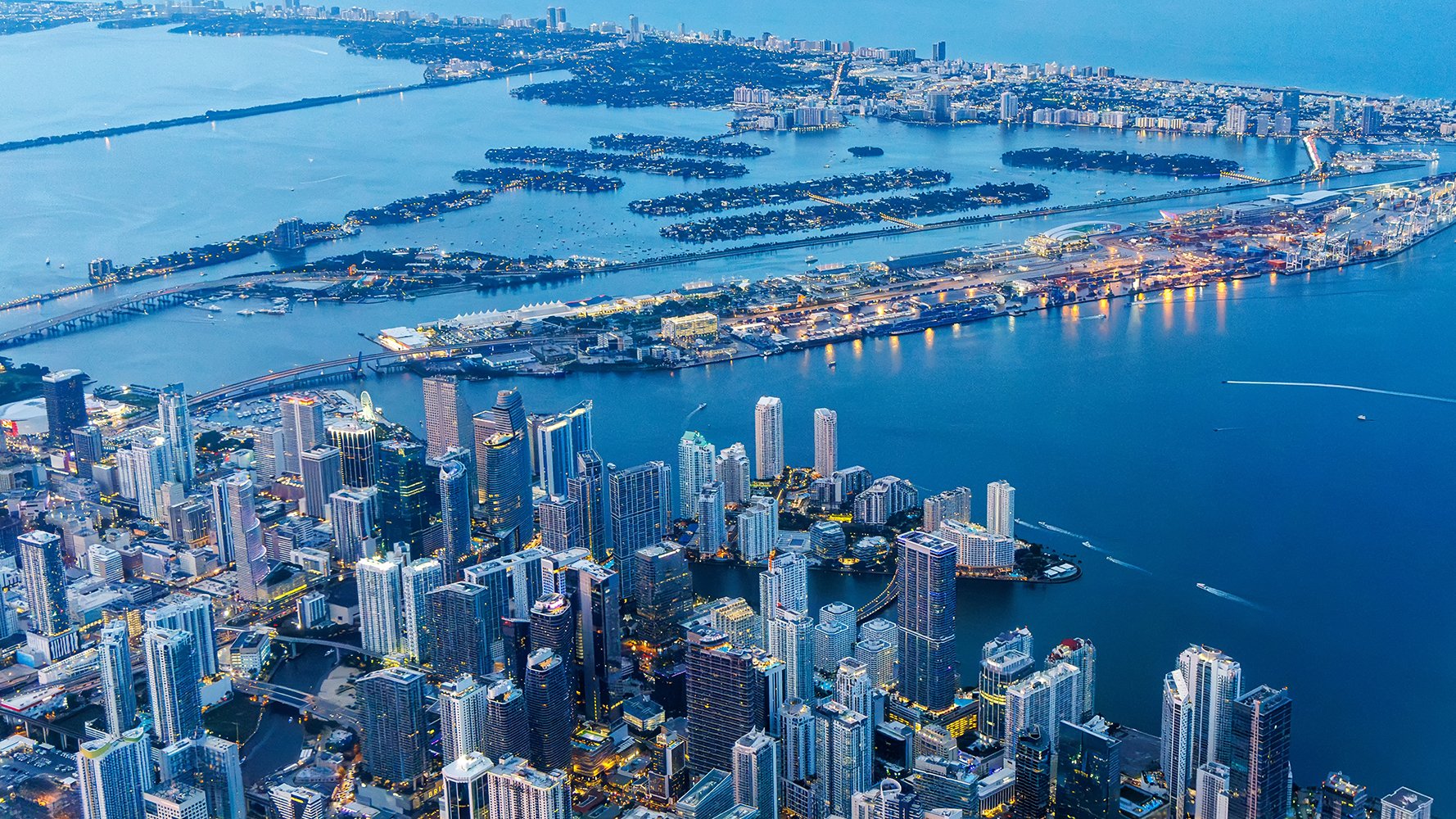 Nighttime aerial view of Miami skyline and Biscayne Bay showcasing luxury high-rise condos and waterfront living. Featuring cityscape.