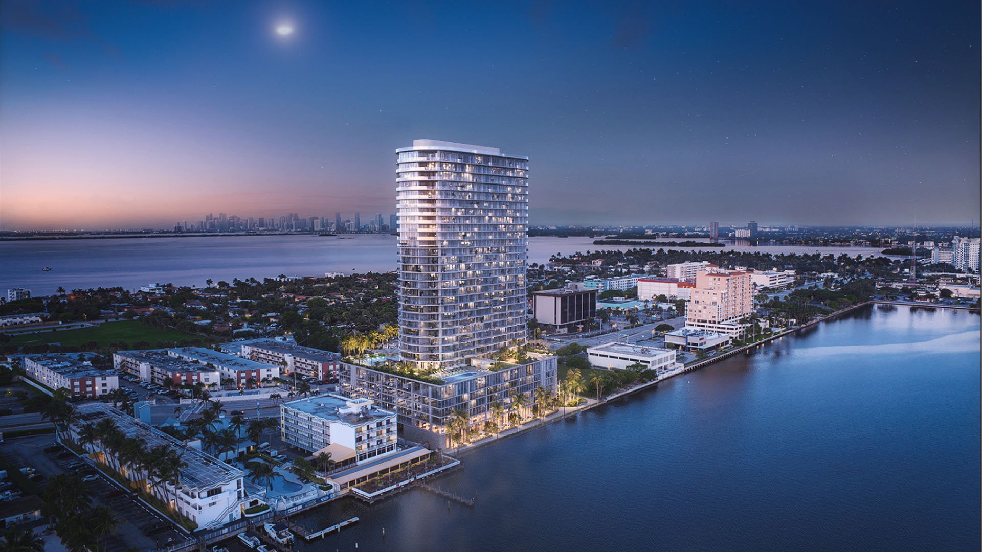Aerial waterfront view of the curved tower and distant skyline at Continuum Club and Residences in North Bay Village, a preconstruction luxury and ultra luxury condos development set along the bay at twilight.