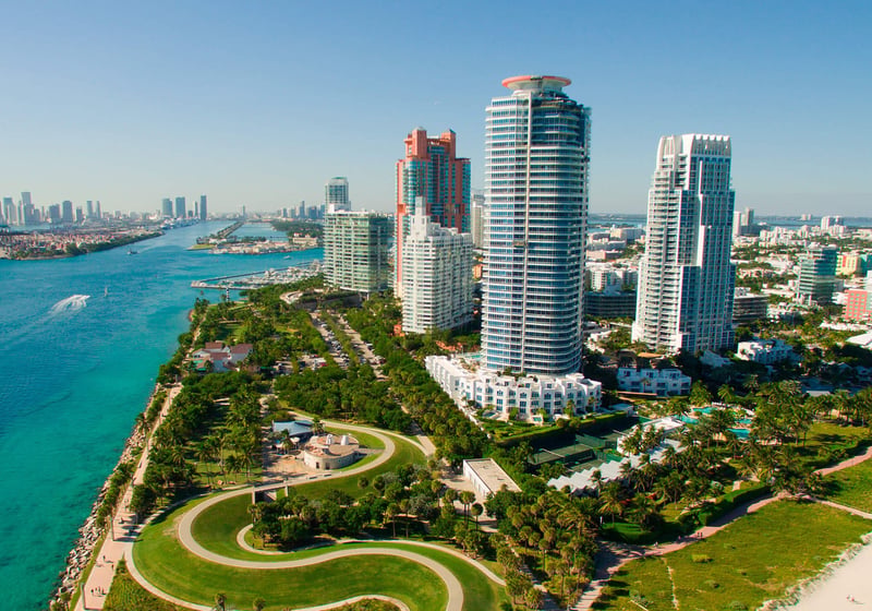 Aerial waterfront view of Continuum on South Beach, Miami Beach, Florida, with luxury and ultra luxury condos beside a sweeping coastal park, turquoise inlet water, and the surrounding skyline.