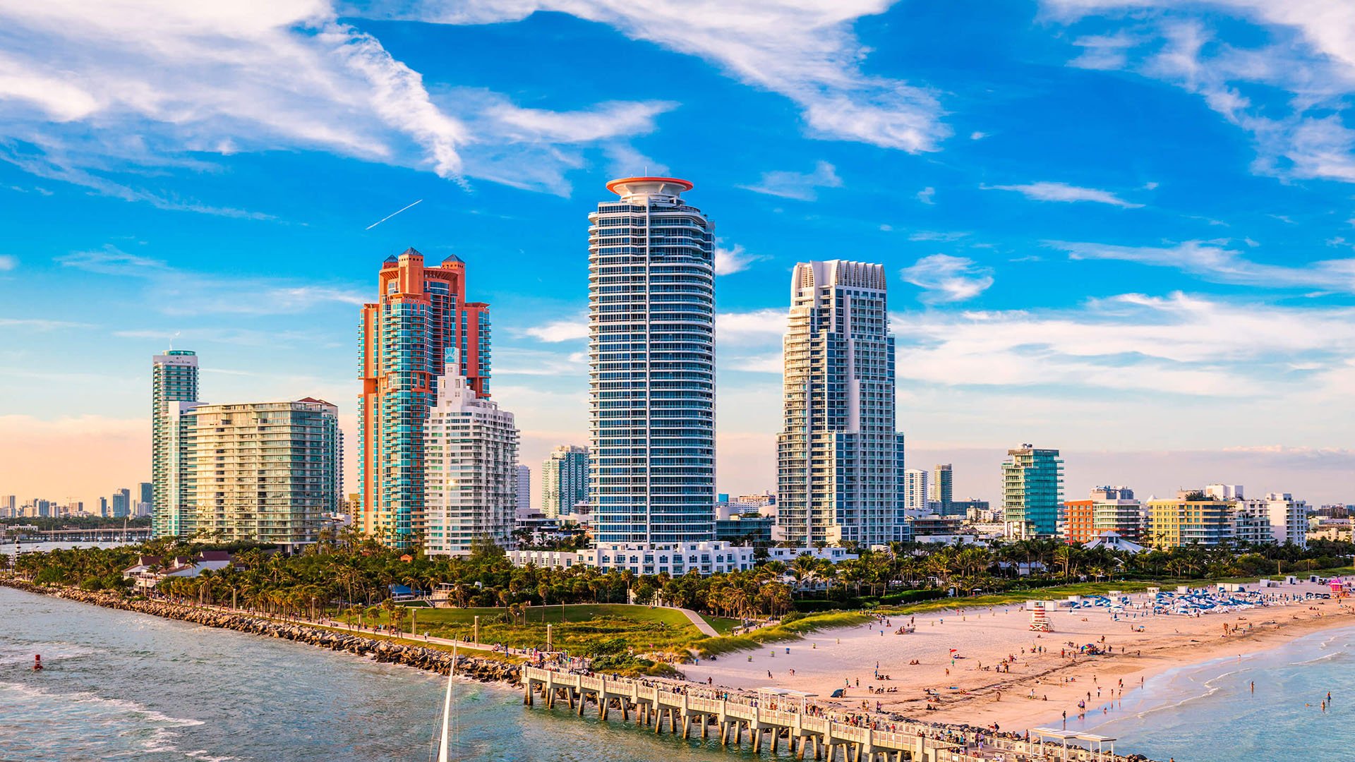 Beachfront skyline view of Continuum on South Beach, Miami Beach, Florida, featuring luxury and ultra luxury condos rising above the sand, jetty, and pier with bright blue sky and ocean water in the foreground.