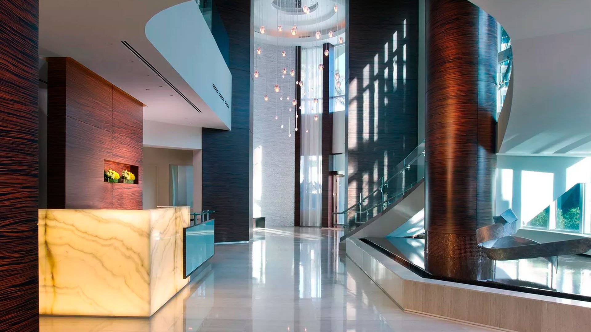 Another view of the double-height lobby at Continuum on South Beach, Miami Beach, Florida, showing luxury and ultra luxury condos with a curved staircase, pendant lights, sunlight, and reflective stone floors.