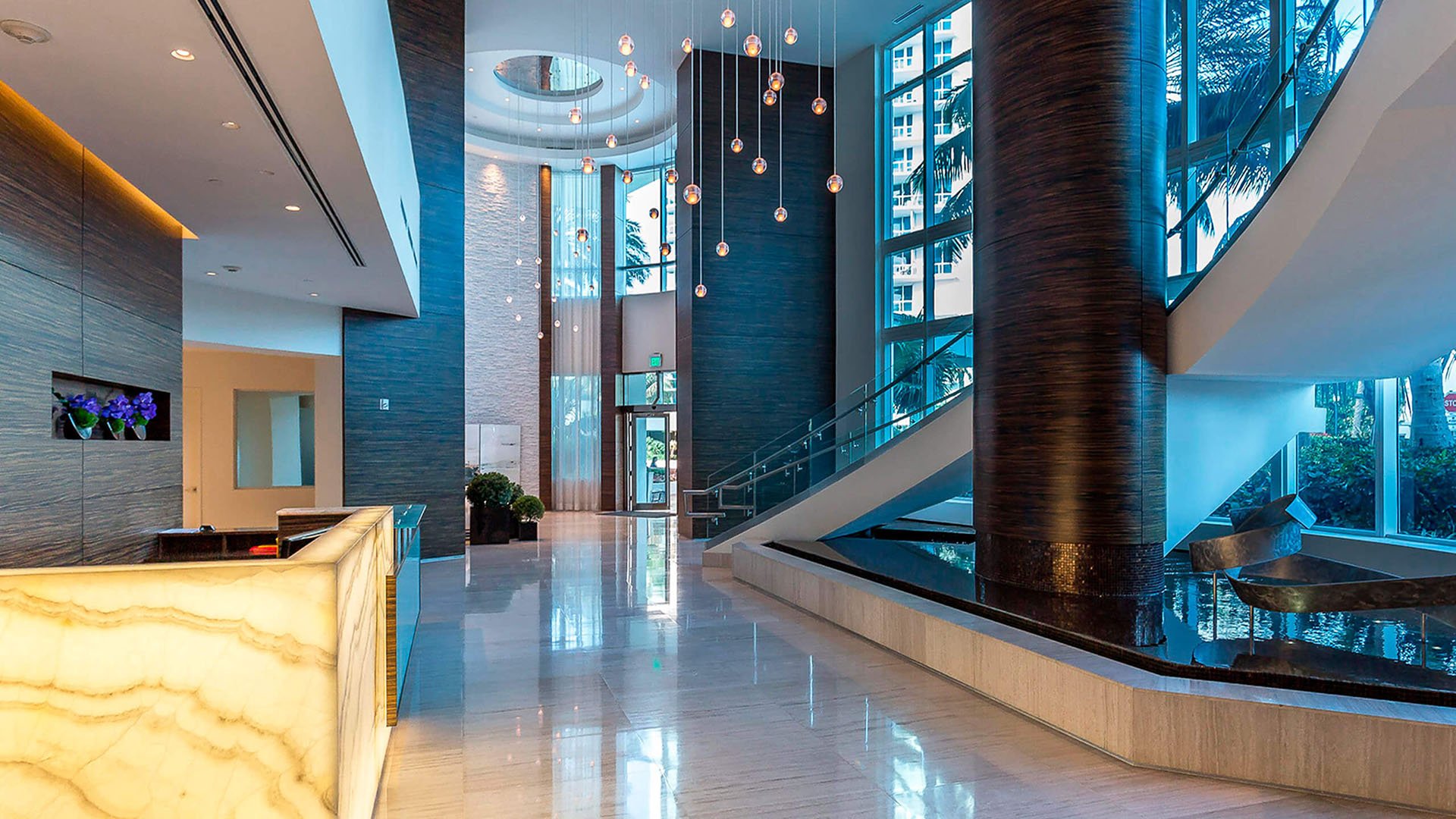 Double-height lobby at Continuum on South Beach, Miami Beach, Florida, showcasing luxury and ultra luxury condos with a glowing reception desk, water feature, sculptural staircase, and dramatic pendant lighting.