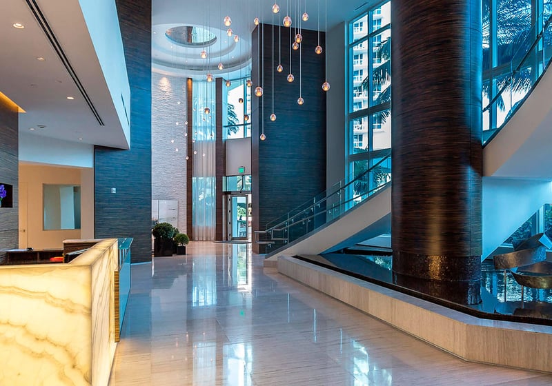 Double-height lobby at Continuum on South Beach, Miami Beach, Florida, showcasing luxury and ultra luxury condos with a glowing reception desk, water feature, sculptural staircase, and dramatic pendant lighting.