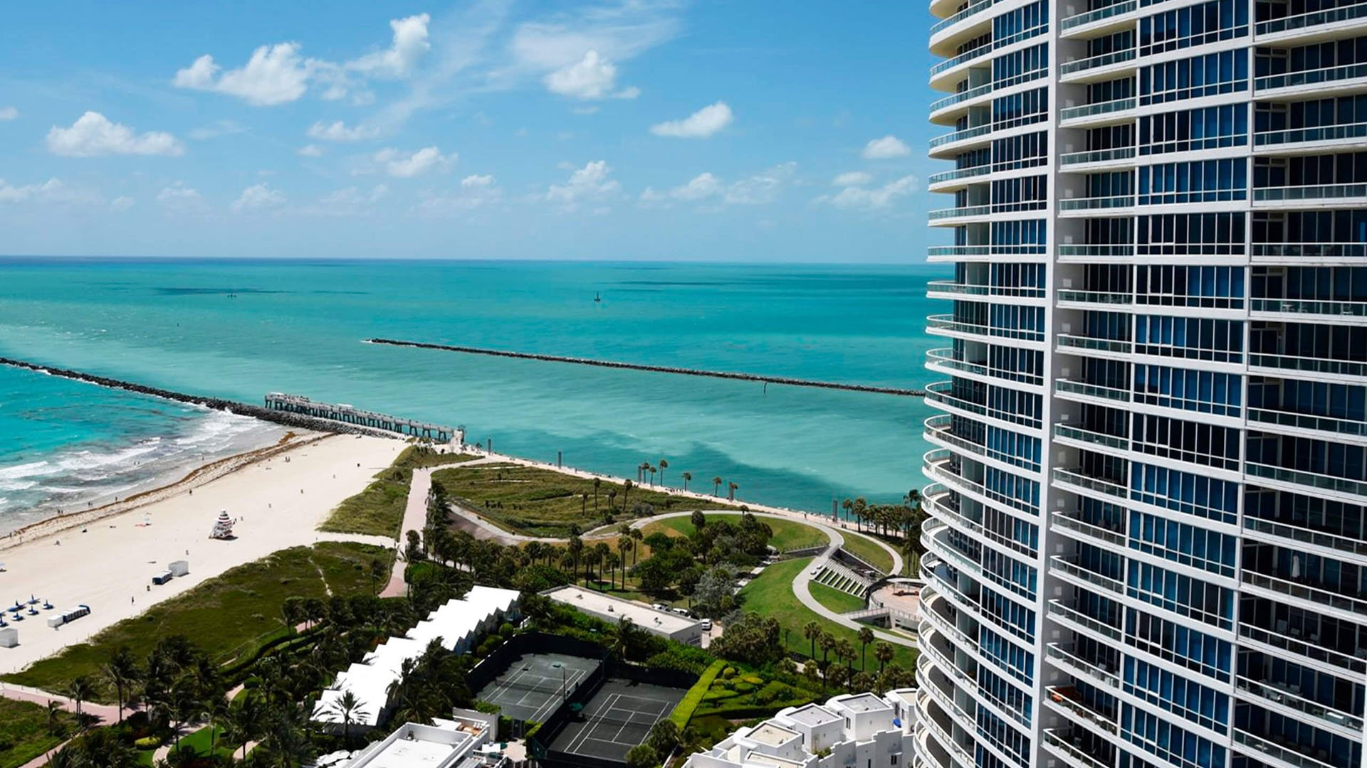 Elevated oceanfront view at Continuum on South Beach, Miami Beach, Florida, featuring luxury and ultra luxury condos beside turquoise water, the sandy shoreline, tennis courts, and the curved tower facade.