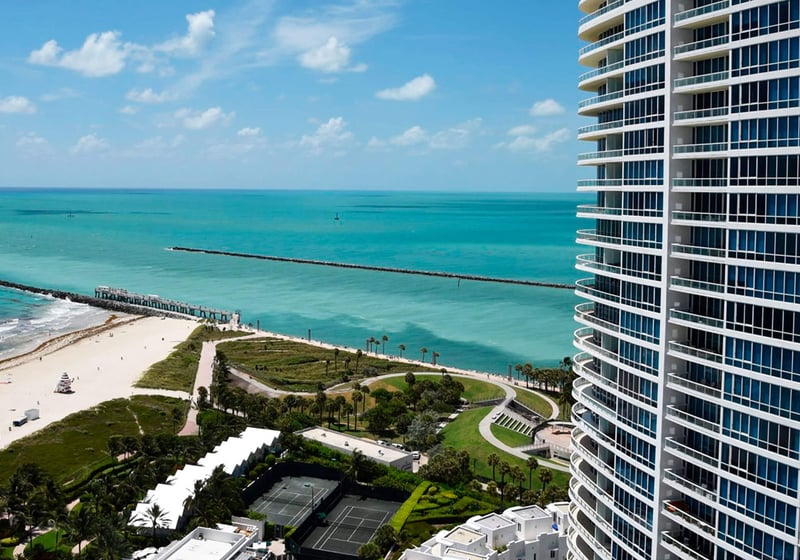 Elevated oceanfront view at Continuum on South Beach, Miami Beach, Florida, featuring luxury and ultra luxury condos beside turquoise water, the sandy shoreline, tennis courts, and the curved tower facade.