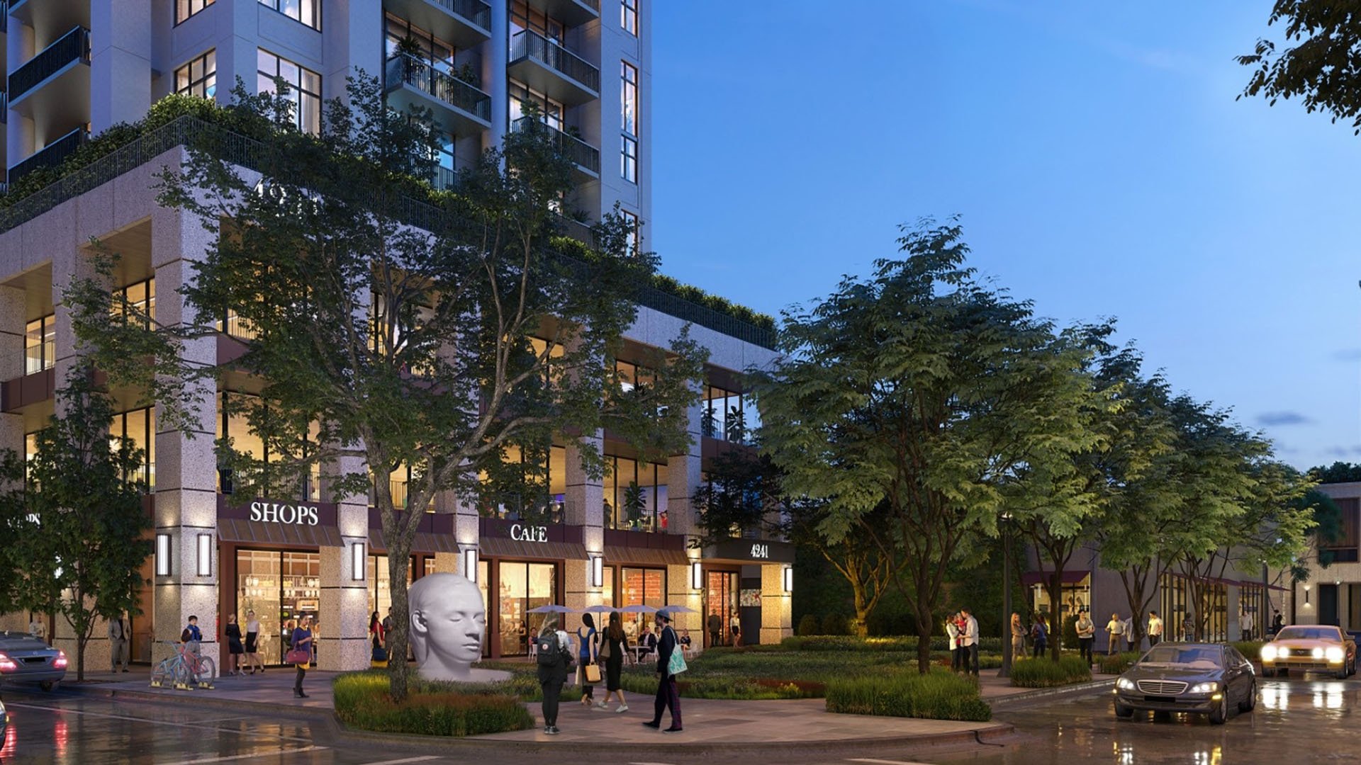 Evening view of the retail plaza at Cora Merrick Park in Coral Gables with illuminated storefronts, a sculptural head, and lush landscaping below luxury and ultra luxury condos.