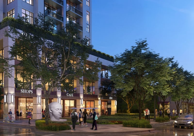 Evening view of the retail plaza at Cora Merrick Park in Coral Gables with illuminated storefronts, a sculptural head, and lush landscaping below luxury and ultra luxury condos.