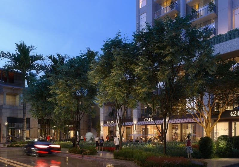Twilight retail courtyard at Cora Merrick Park in Coral Gables with glowing storefronts, mature trees, and passing cars beneath luxury and ultra luxury condos.