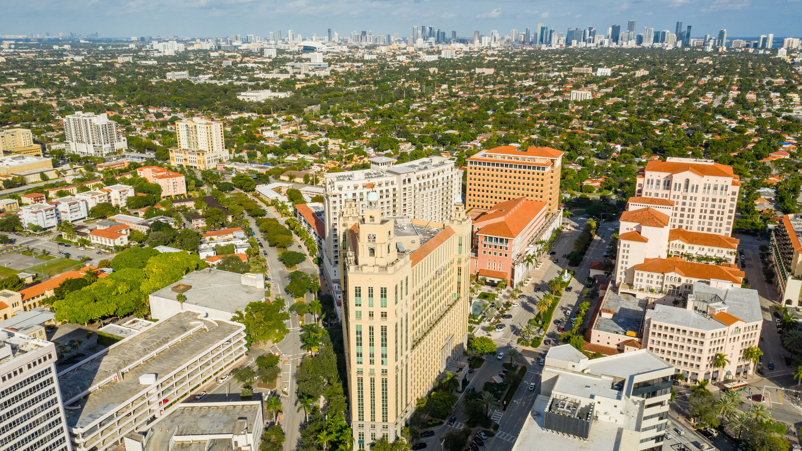 Aerial of Coral Gables with Miami Downtown beyond, tree‑lined neighborhoods linking to luxury and ultra luxury condos, preconstruction and resale. Featuring cityscape and view.