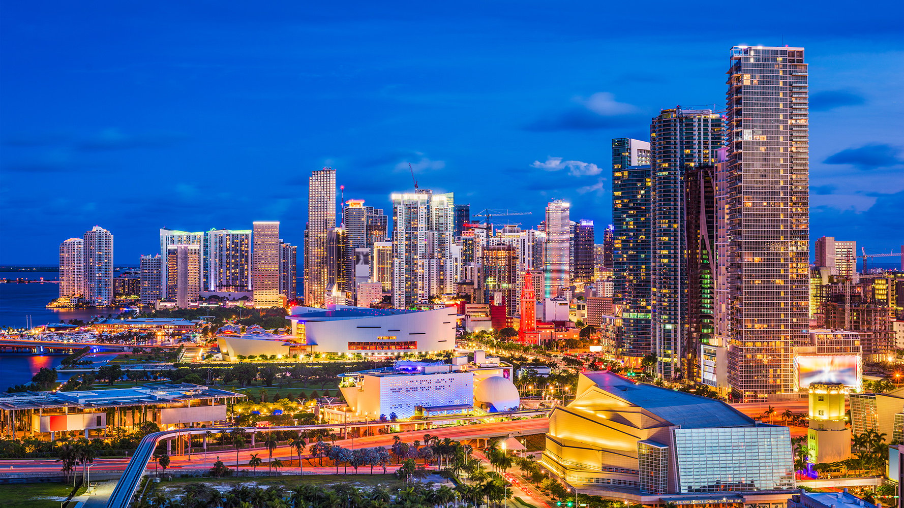 Downtown Miami night skyline glowing over Biscayne Bay, iconic towers and waterfront lights; prime market for luxury and ultra luxury condos, with preconstruction and resale inventory. Featuring view.