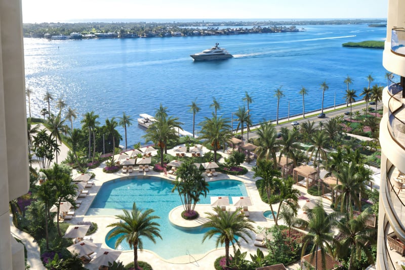 Edgeworth West Palm Beach luxury ultra luxury condos amenity deck overlooking the waterfront, with a resort-style pool, palm-lined terraces, lounge seating, and a marina view with a yacht passing by.