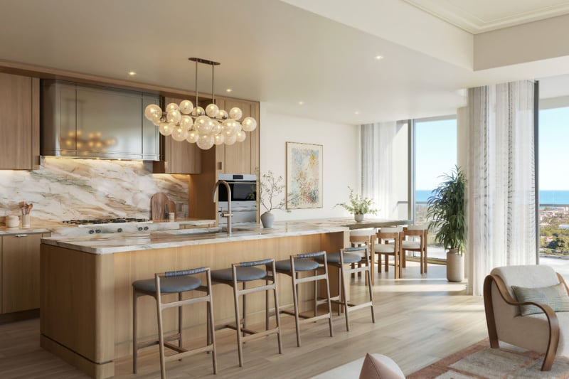 Edgeworth West Palm Beach luxury ultra luxury condos chef kitchen with a large marble island, warm wood cabinetry, pendant lighting, and open-plan dining space beside expansive windows with water views.