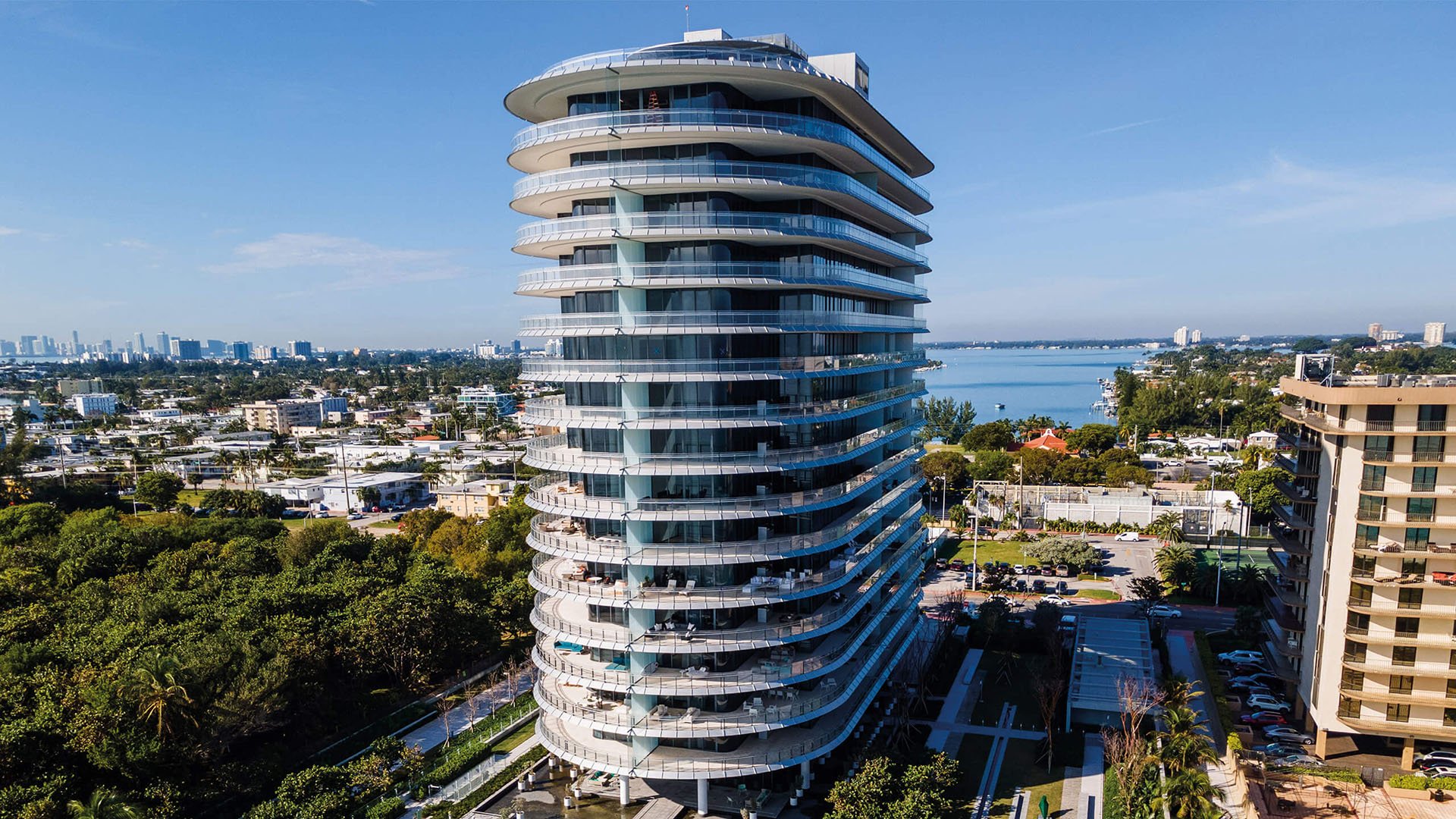 Eighty Seven Park, Miami Beach luxury and ultra luxury condos tower rising above tropical grounds with a calm bay backdrop and city skyline in the distance.