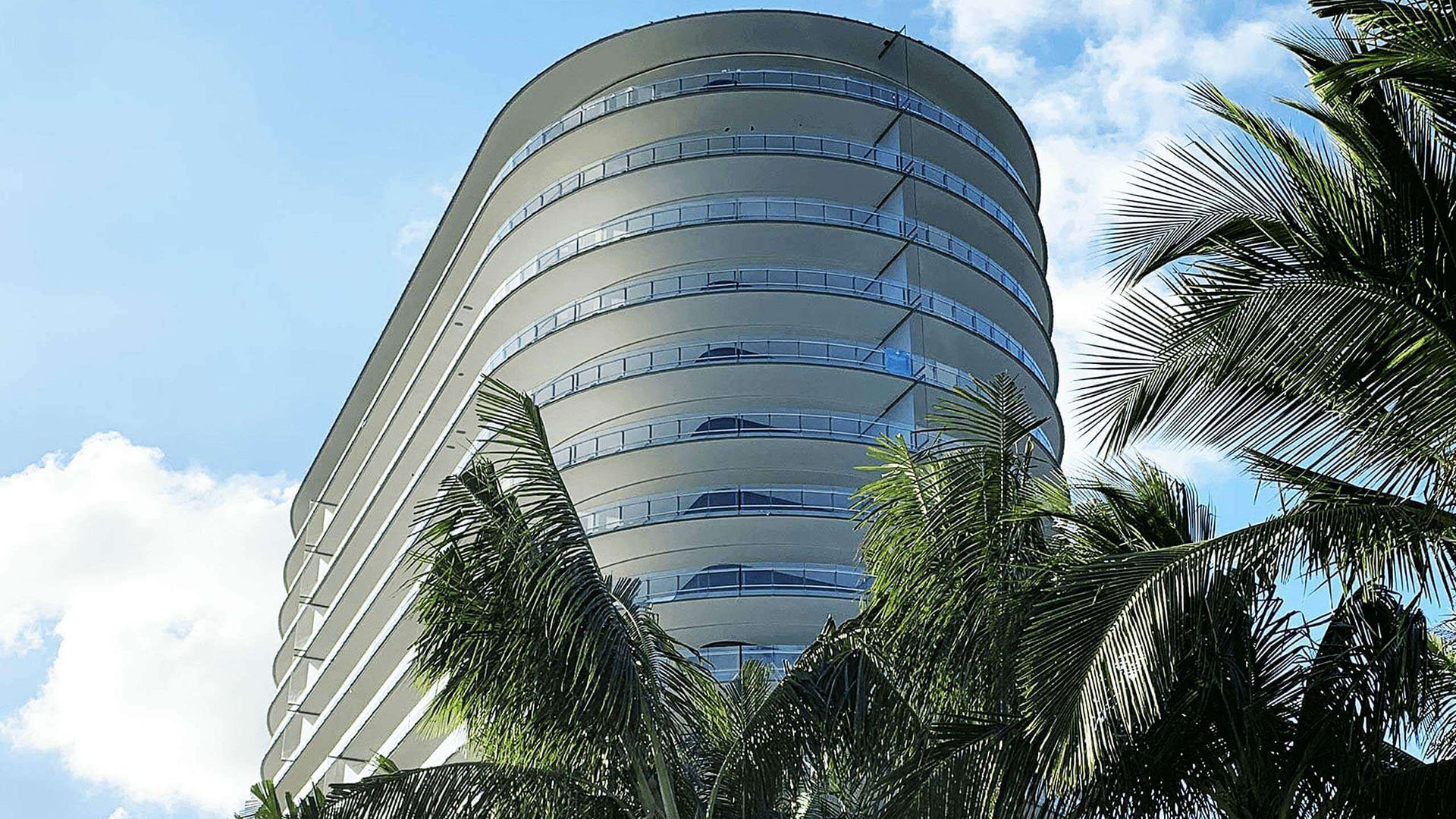 Eighty Seven Park, Miami Beach luxury and ultra luxury condos exterior looking up at the curved oceanfront tower framed by tall palm fronds and blue sky.