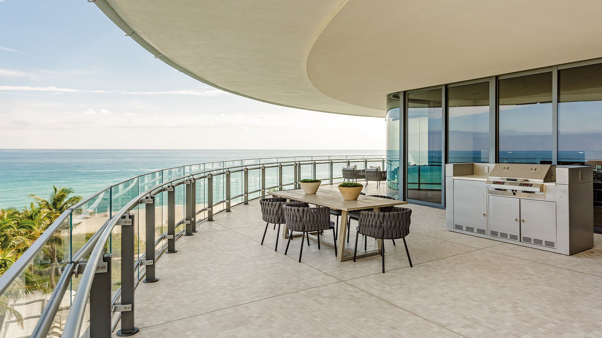 Eighty Seven Park, Miami Beach luxury and ultra luxury condos curved terrace with glass railings, outdoor dining, a built-in grill, and sweeping ocean views.