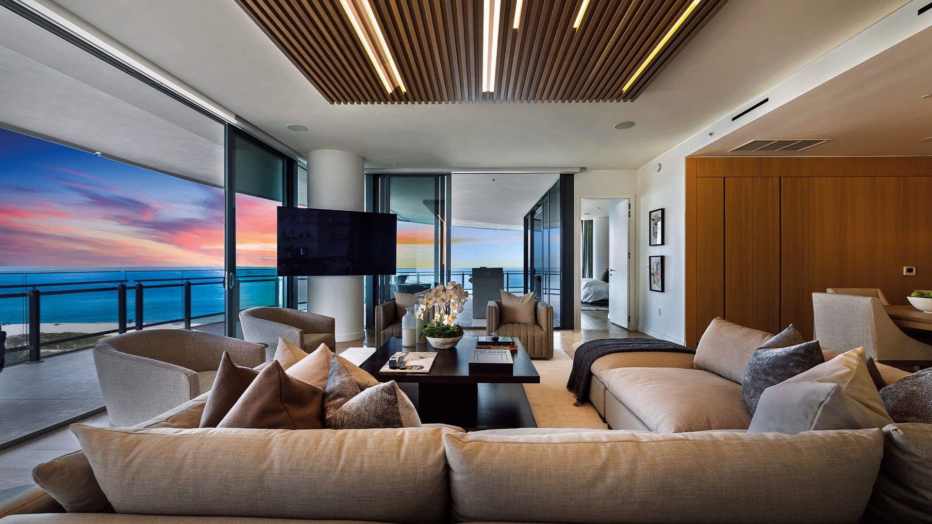 Eighty Seven Park, Miami Beach luxury and ultra luxury condos living room with plush seating, a slatted ceiling, large television, and a sunset ocean view from the terrace.