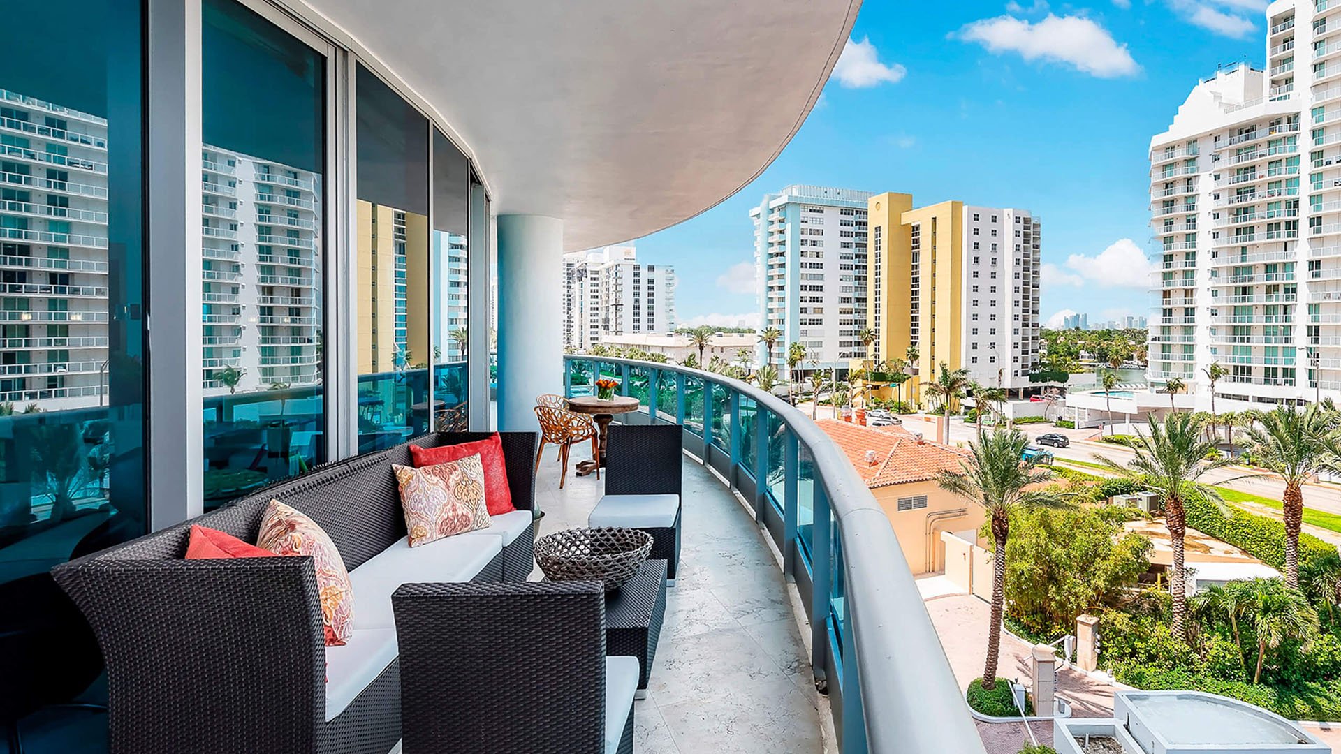 Curved balcony lounge at Faena House in Miami Beach, luxury and ultra luxury condos with outdoor seating, blue glass railings, palm trees below, and a neighboring skyline view.