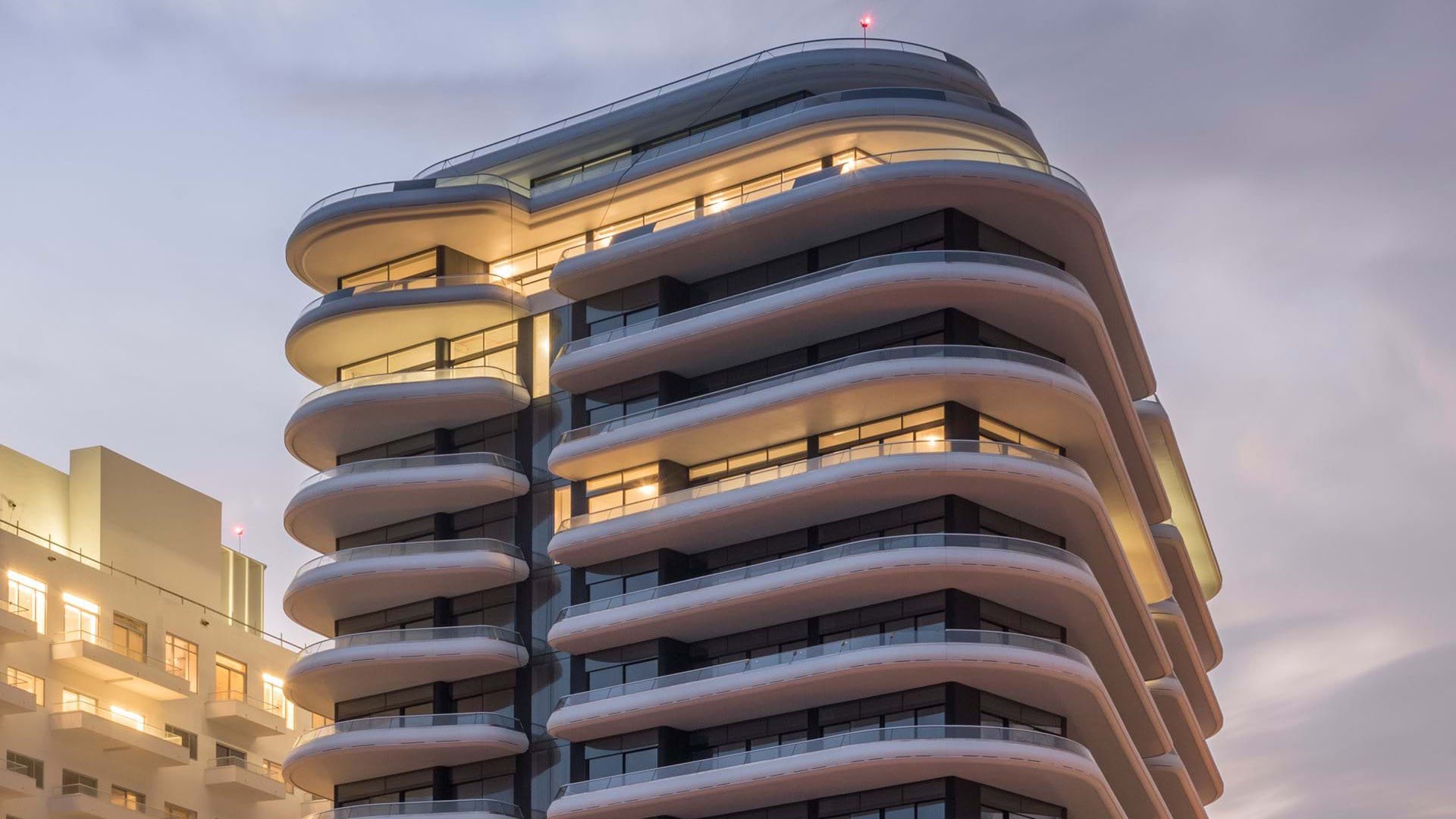 Upward dusk view of Faena House in Miami Beach, luxury and ultra luxury condos with illuminated curved balconies, smooth white bands, dark glass, and neighboring buildings.