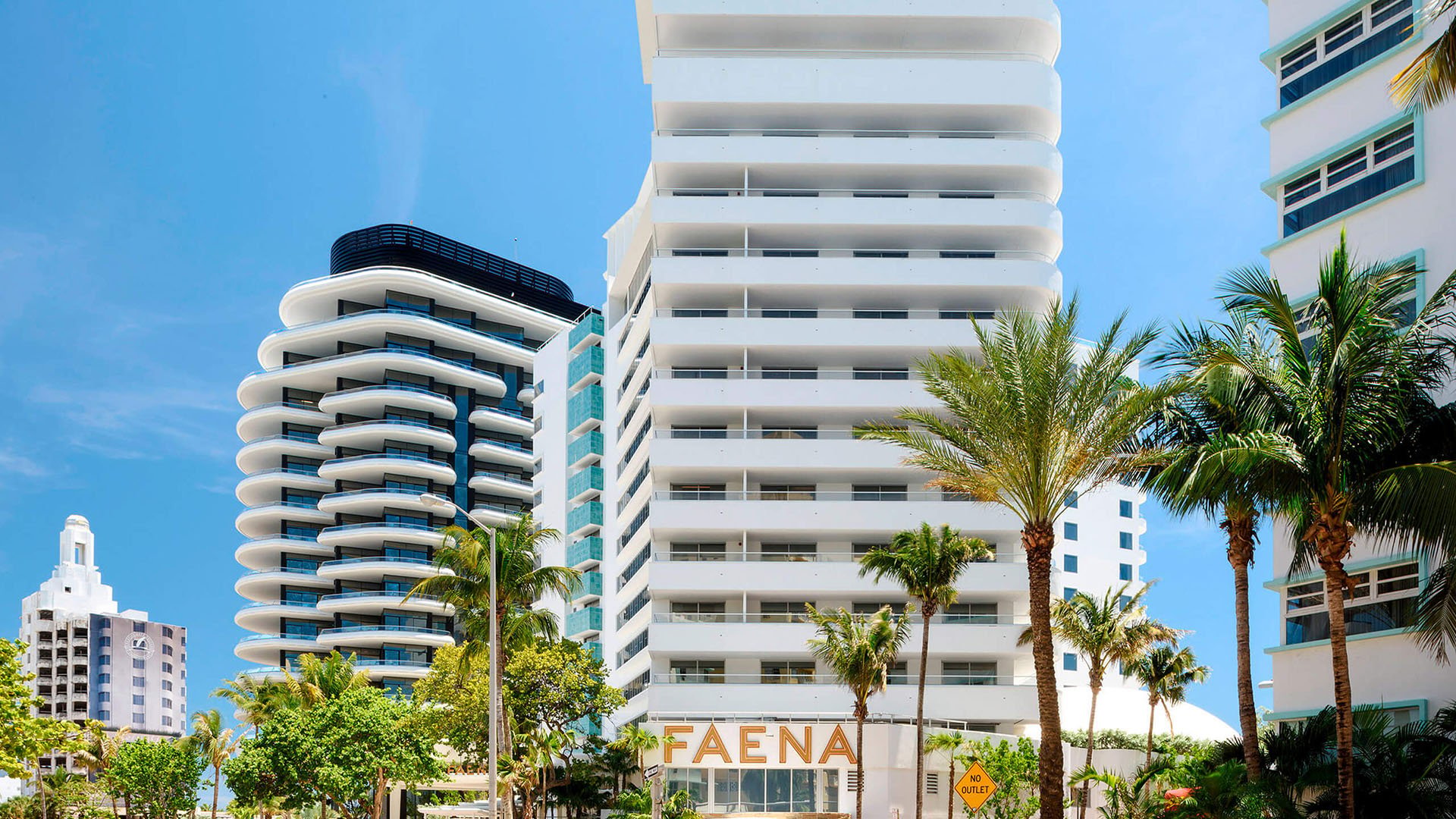 Street-level exterior of Faena House in Miami Beach, luxury and ultra luxury condos with the Faena sign, curved balconies, white neighboring towers, and palm-lined streets.
