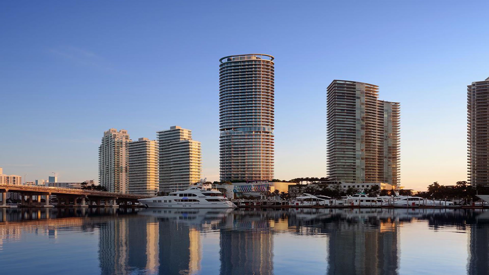Waterfront view of the Five Park tower in Miami Beach, luxury and ultra luxury condos reflected beside a marina and skyline at dusk.