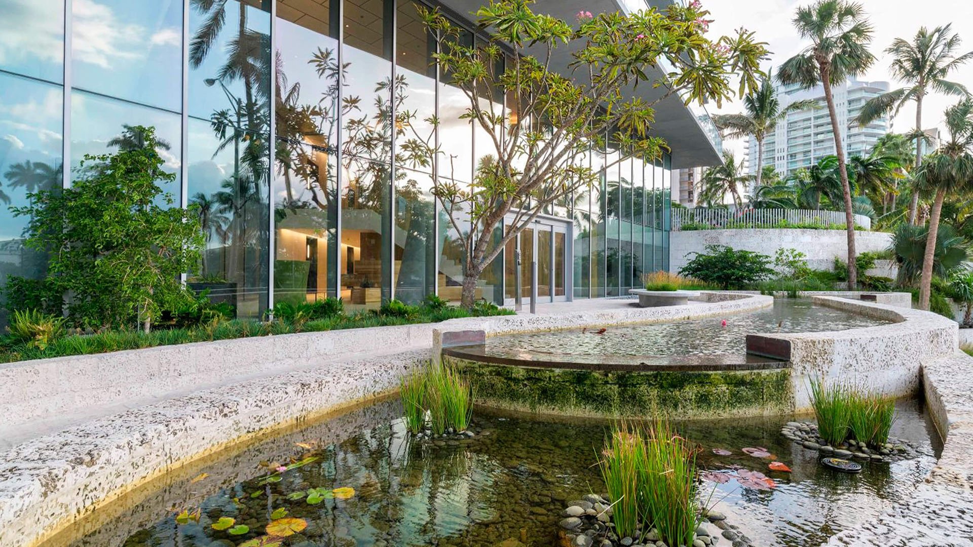 Grove at Grand Bay, Coconut Grove luxury and ultra luxury condos with a glass entrance facade, reflecting pond, curved stone edges, and tropical landscaping.