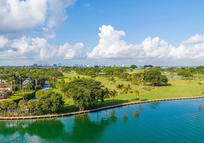 Indian Creek Residences and Yacht Club Bay Harbor Islands, Miami, Florida aerial showing waterfront golf course greens and tree-lined fairways with city skyline, highlighting the setting for luxury and ultra luxury preconstruction condos.