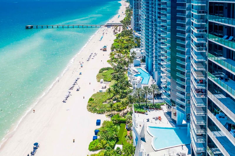 Aerial beachfront skyline view of Jade Ocean in Sunny Isles Beach, showing luxury and ultra luxury condos along turquoise water with a long pier, sandy shoreline, and neighboring oceanfront towers.