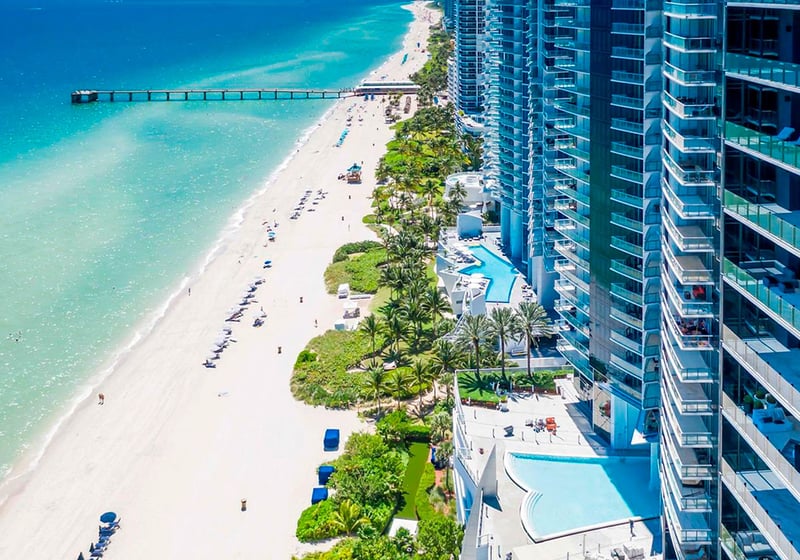 Aerial beachfront skyline view of Jade Ocean in Sunny Isles Beach, showing luxury and ultra luxury condos along turquoise water with a long pier, sandy shoreline, and neighboring oceanfront towers.