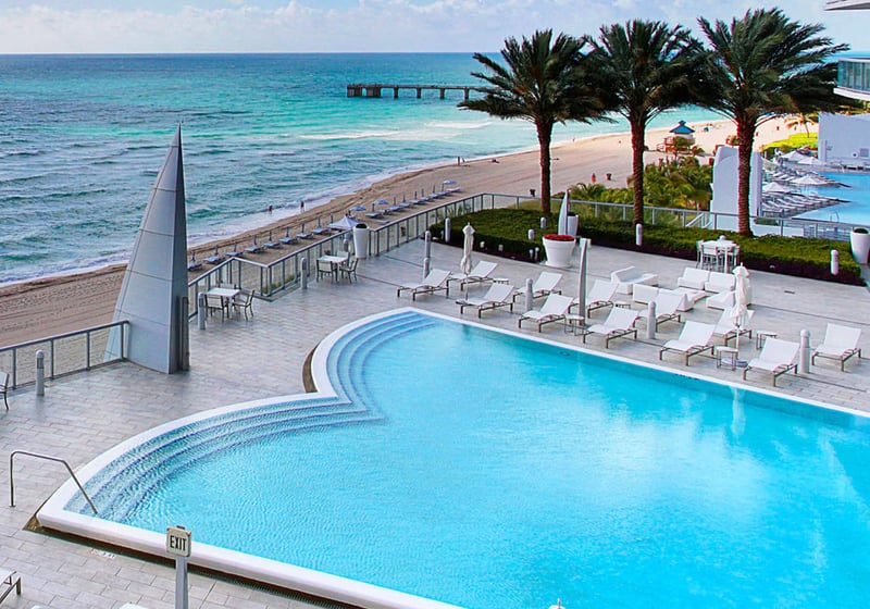 Beachfront pool terrace at Jade Ocean in Sunny Isles Beach, where luxury and ultra luxury condos overlook a curved oceanfront pool, palm-lined sundeck, white loungers, and the beach.